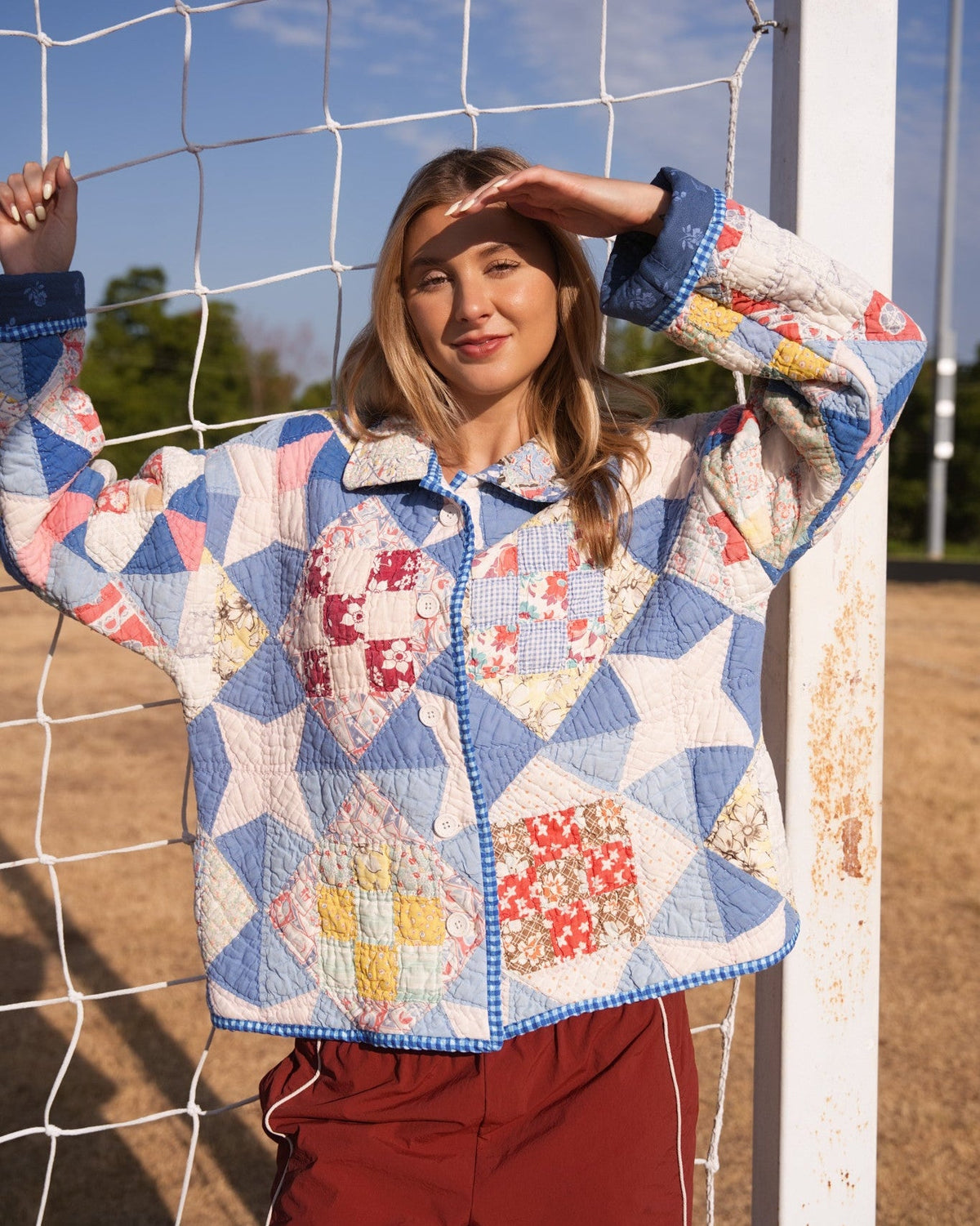 Person sanding outdoors in front of a white soccer net, wearing a quilted patchwork jacket with blue trim and red-brown pants.