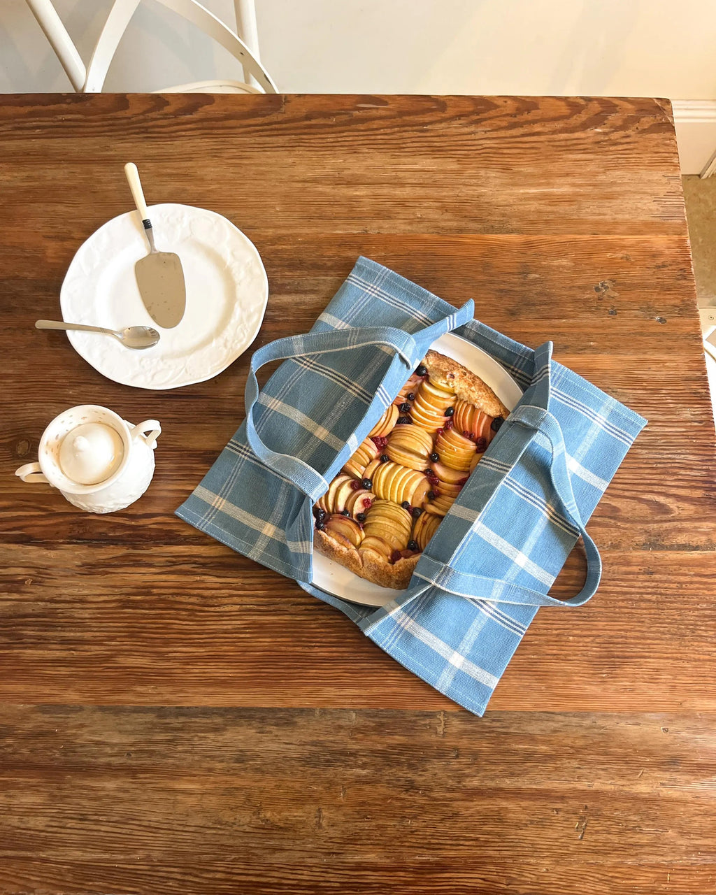 A blue and white plaid reusable pie carrier tote bag filled with a pie sits on a rustic wooden table, styled with a white plate, spoon, and a small cup of coffee.