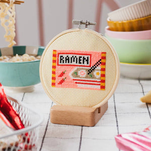 Cross-stitched 'RAMEN' design in a wooden hoop with bowls and noodles in the background.