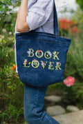 Person holding a blue denim tote bag with 'BOOK LOVER' embroidery in an outdoor setting.