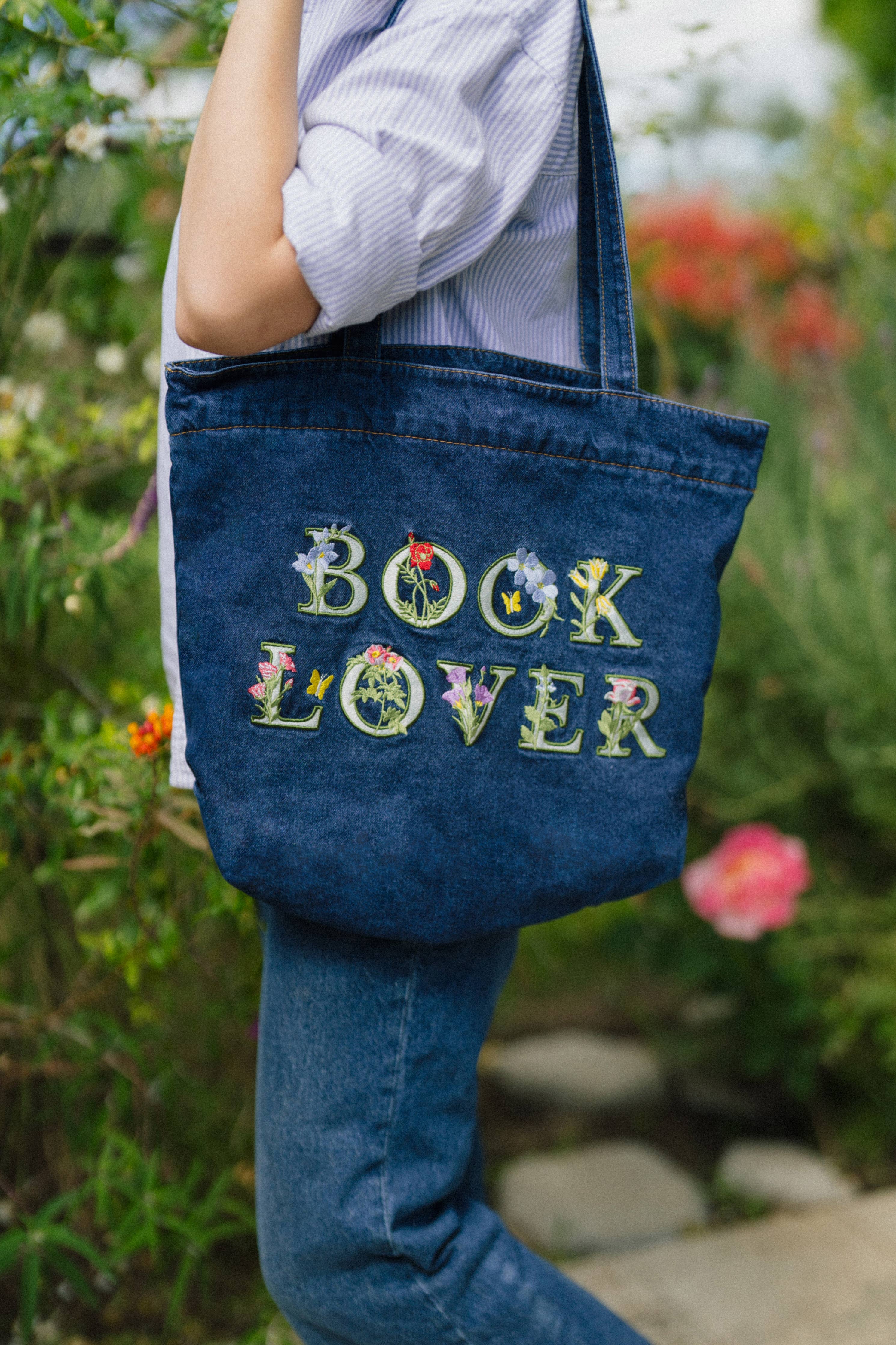 Person holding a blue denim tote bag with 'BOOK LOVER' embroidery in an outdoor setting.