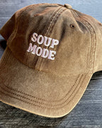 Brown cap with 'SOUP MODE' text on a wooden surface