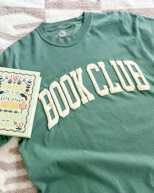 Green t-shirt with 'BOOK CLUB' text and a book on a striped fabric background