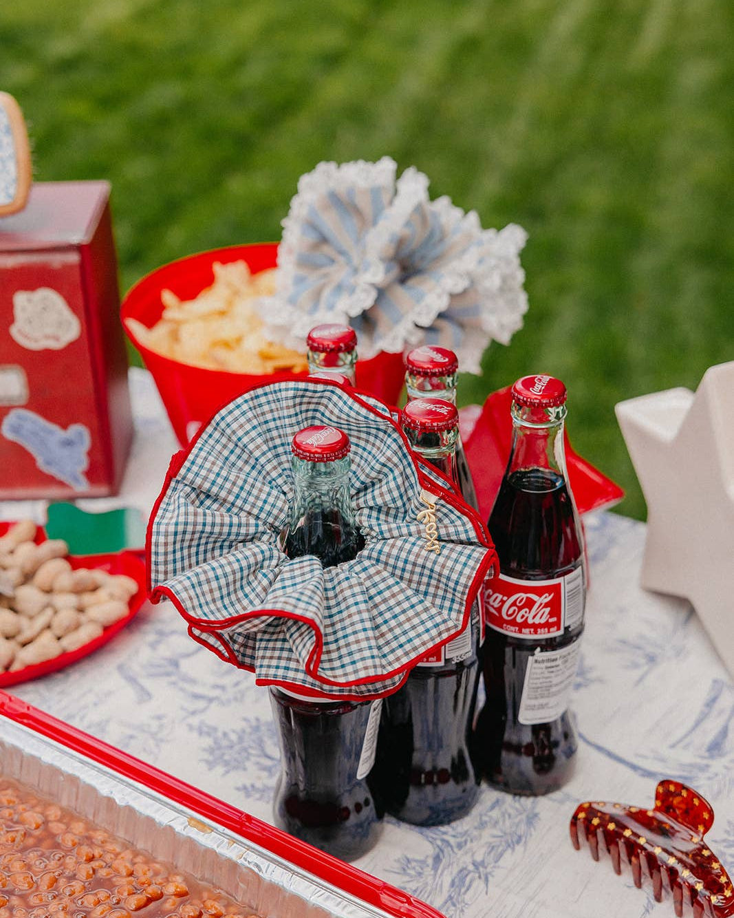 A styled outdoor picnic with Coca-Cola glass bottles, and blue-and-white scrunchie with a red trim, with a blurred green lawn in the background.