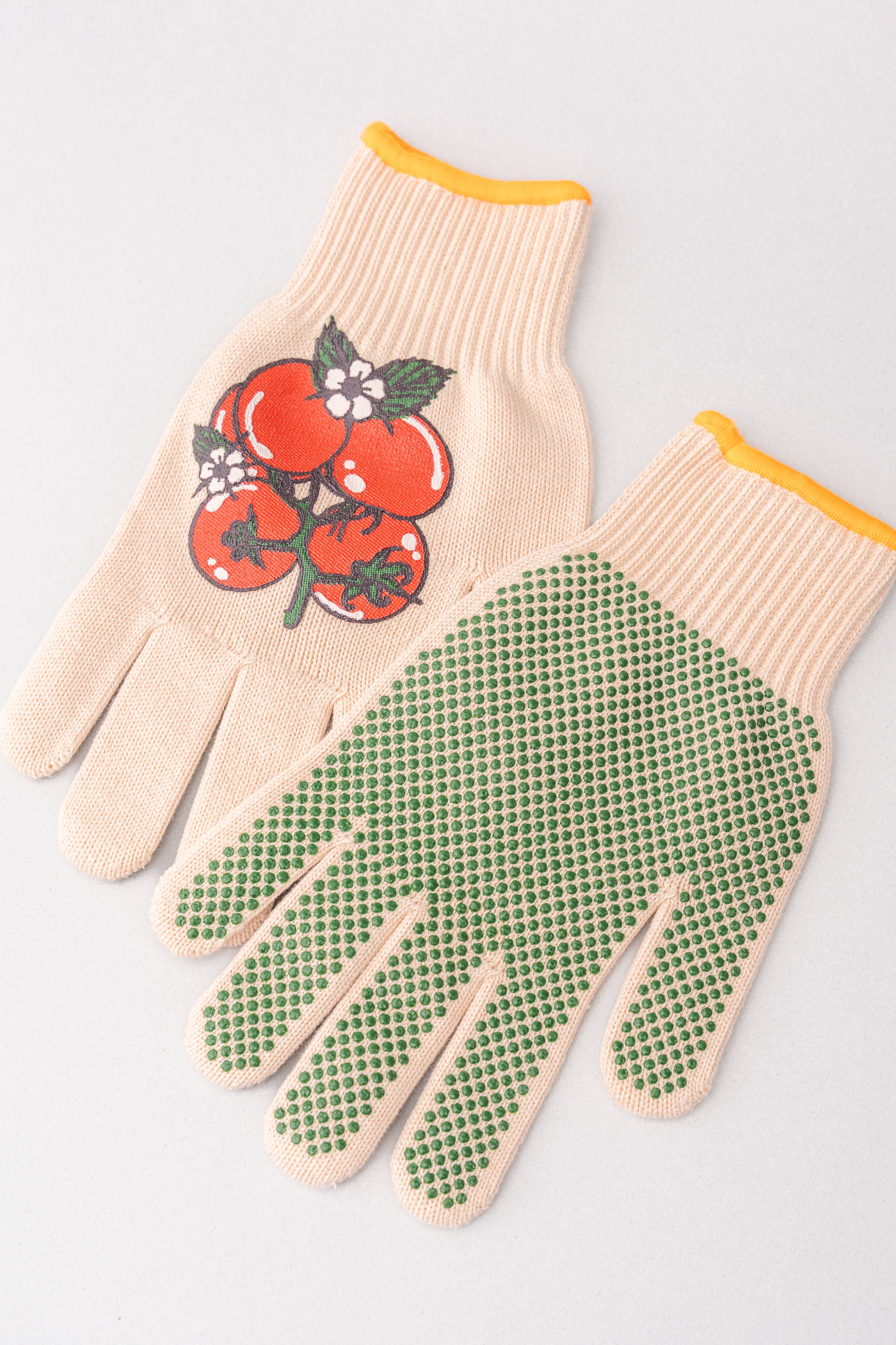 A pair of knit gardening gloves on a light gray background. One glove shows a cream-colored knit body with a graphic of red tomatoes and green leaves on the back, finished with a yellow cuff.
