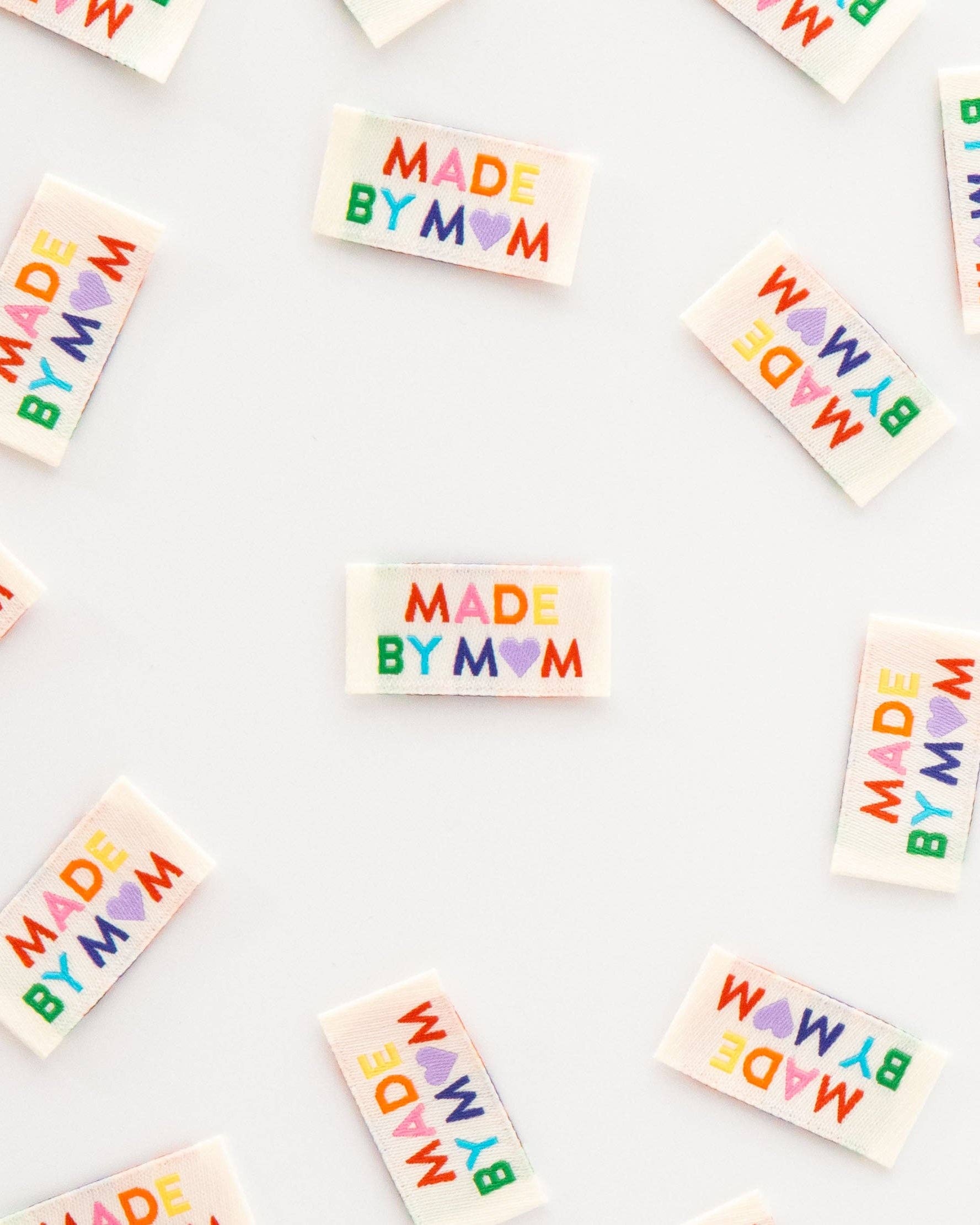 White rectangular sew-in labels with the text 'MADE BY MOM' in colorful rainbow letters and a heart, scattered on a white background.