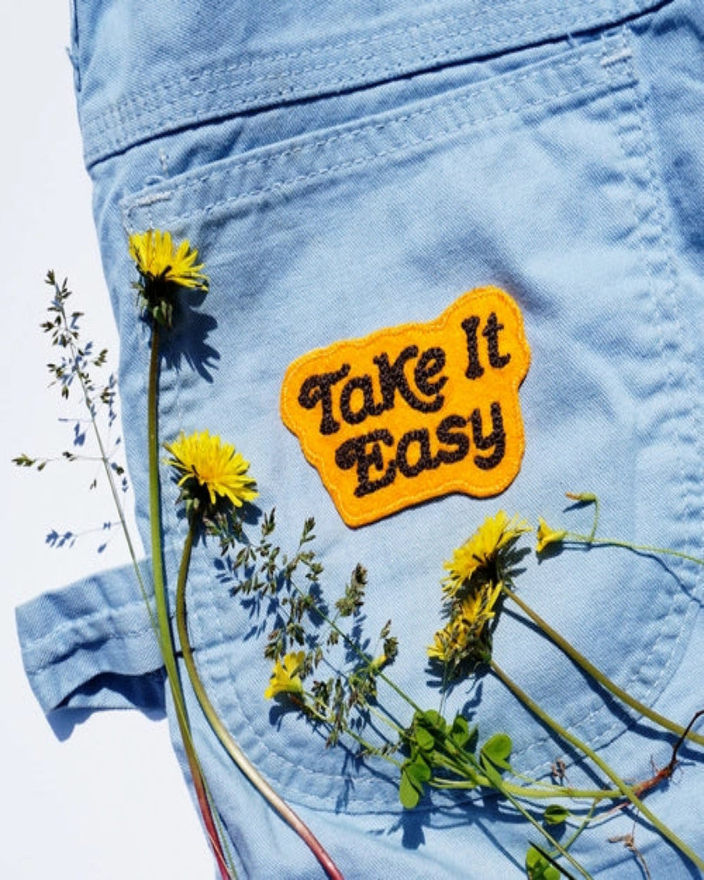Blue denim pant with yellow 'Take It Easy' patch and dandelions on a white background