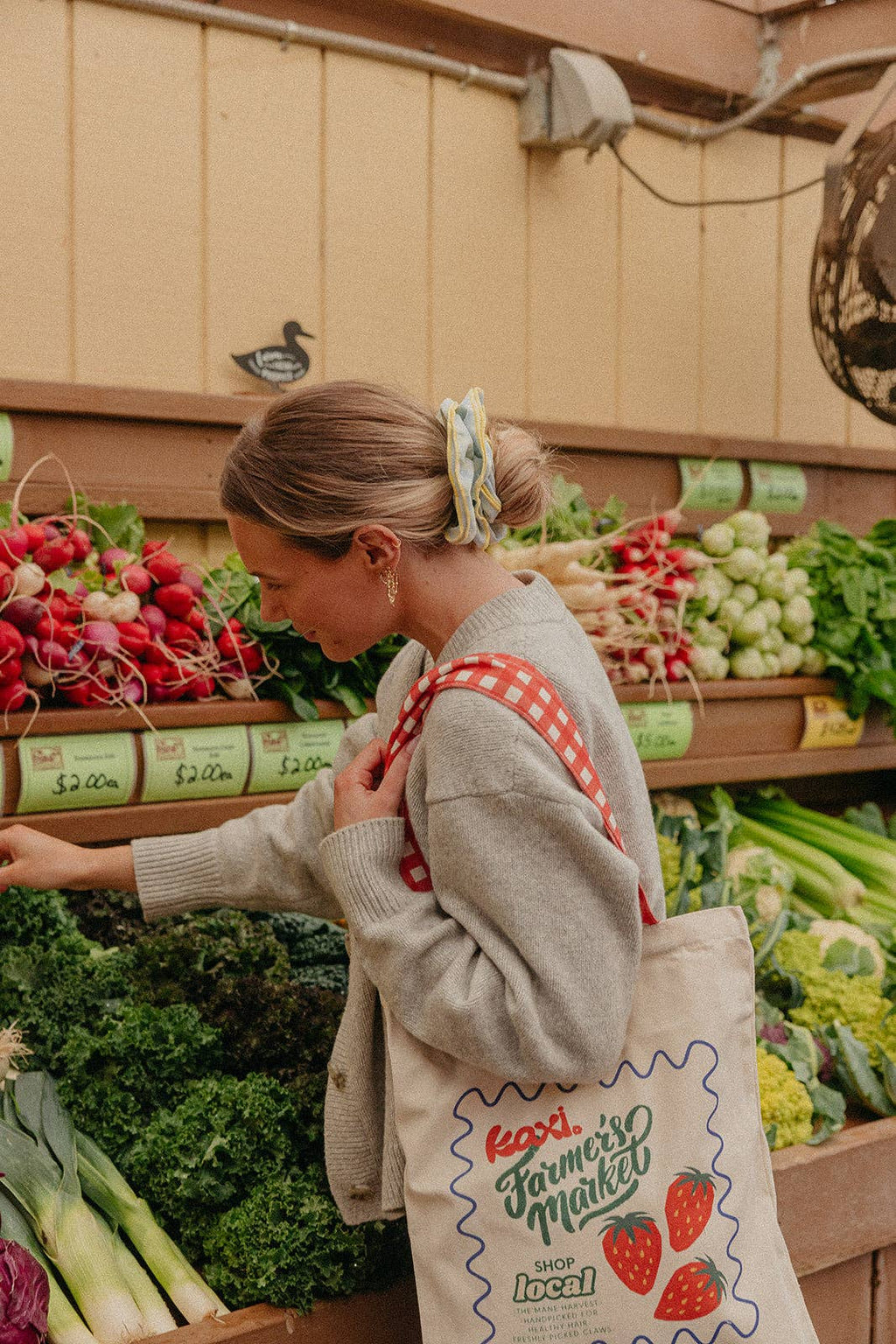 Woman shopping at a farmer's market wearing a blue and yellow scrunchie with a reusable bag.