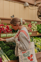 Woman shopping at a farmer's market wearing a blue and yellow scrunchie with a reusable bag.