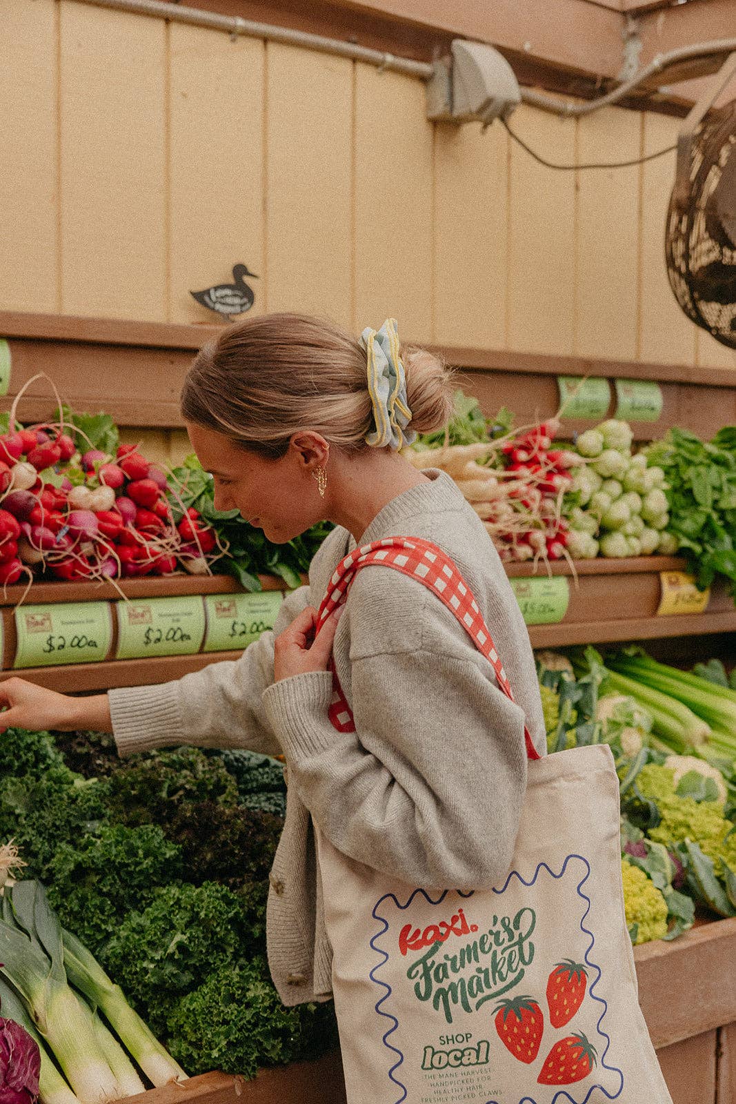 Woman shopping at a farmer's market wearing a blue and yellow scrunchie with a reusable bag.