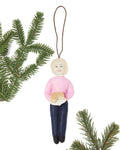A handmade felt Christmas ornament of Martha Stewart pink sweater and blue jeans, holding a cake. The ornament hangs from a brown string against a white background, decorated with green pine branches in the corners.