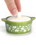A small green ceramic lidded dish inspired trinket box with a white floral pattern on the exterior and a white lid being lifted by a person's fingers, shown on a white background.