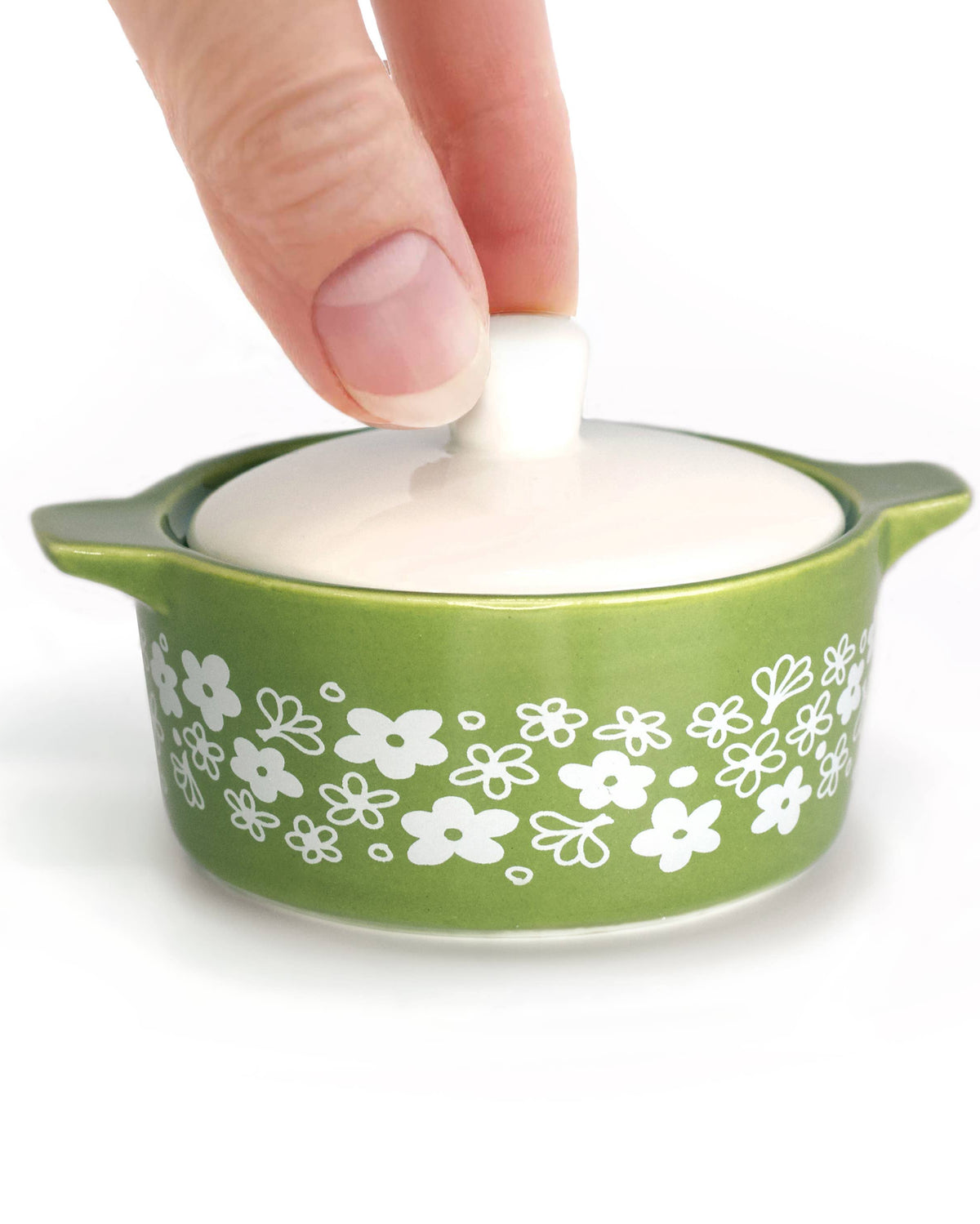 A small green ceramic lidded dish inspired trinket box with a white floral pattern on the exterior and a white lid being lifted by a person's fingers, shown on a white background.