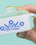 Small white ornament with blue floral design held by a hand against a light blue background