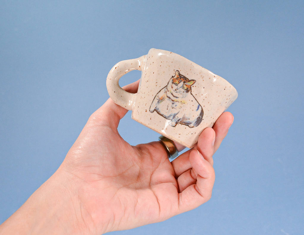 Hand holding a small ceramic mug with a cat design against a blue background