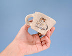 Hand holding a small ceramic mug with a cat design against a blue background