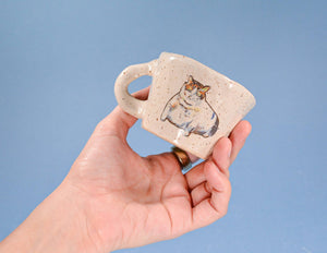 Hand holding a small ceramic mug with a cat design against a blue background