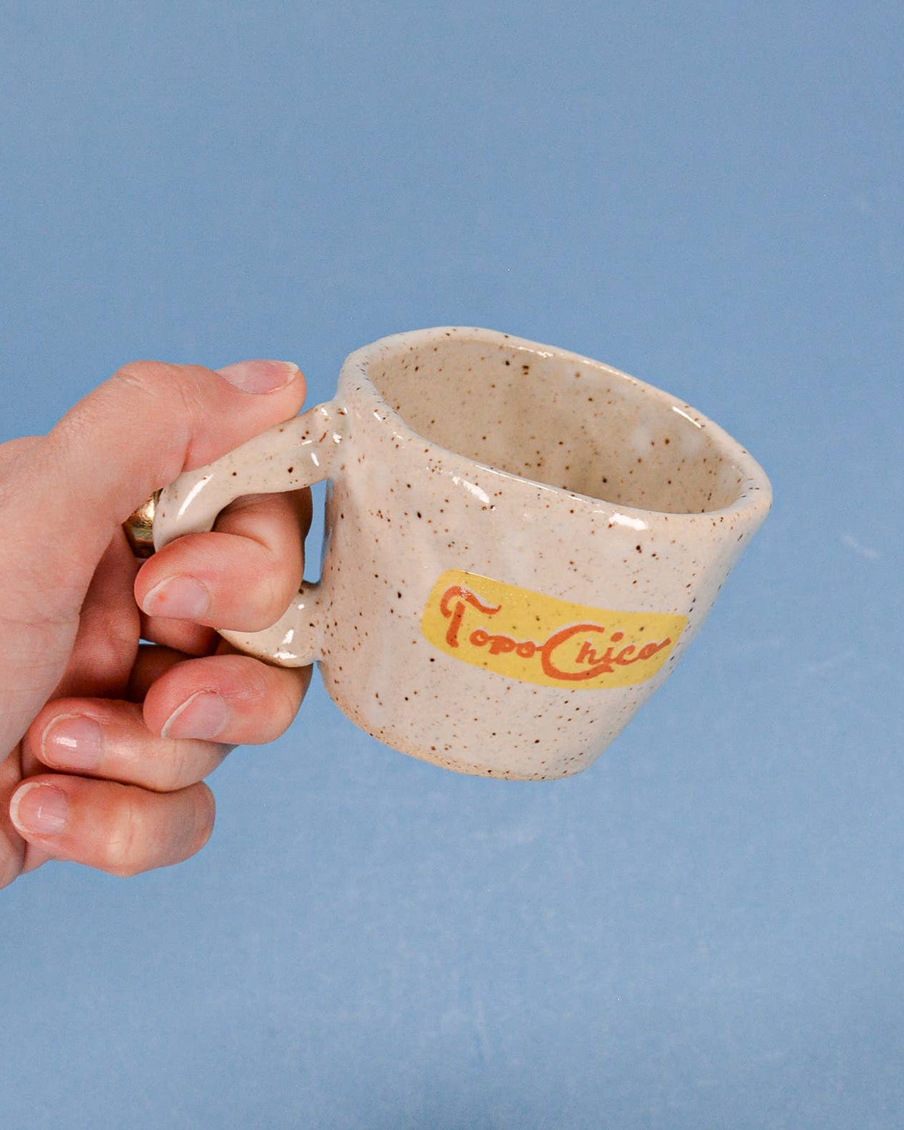 Hand holding a ceramic mug with 'Topo Chico' branding against a blue background