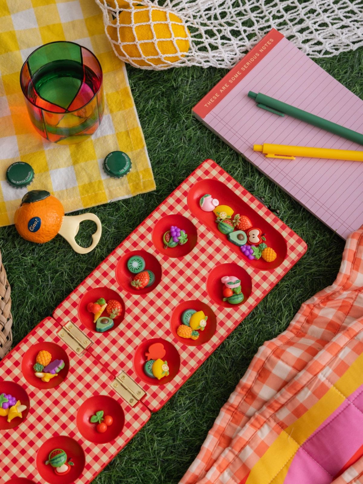 Colorful outdoor setting with a red checkered mat, colorful drink, and stationery items on grass.