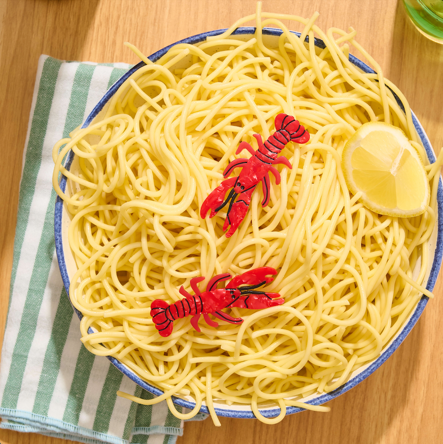 Bowl of spaghetti with plastic lobsters hair clips and a lemon wedge on a wooden table.