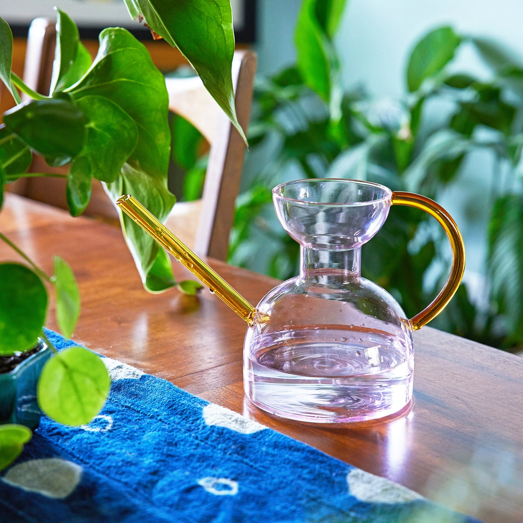 Pink Clear glass watering can with gold handle on a wooden table with plants in the background