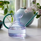 Purple/blue clear glass watering can with green handle on a white surface with plants in the background