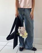 A person wearing loose-fit blue jeans and a pale pink top stands indoors against a white wall, holding a black flower tote with a bouquet of light pink and white flowers in it.
