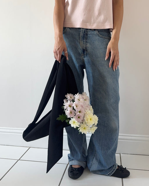 A person wearing loose-fit blue jeans and a pale pink top stands indoors against a white wall, holding a black flower tote with a bouquet of light pink and white flowers in it.