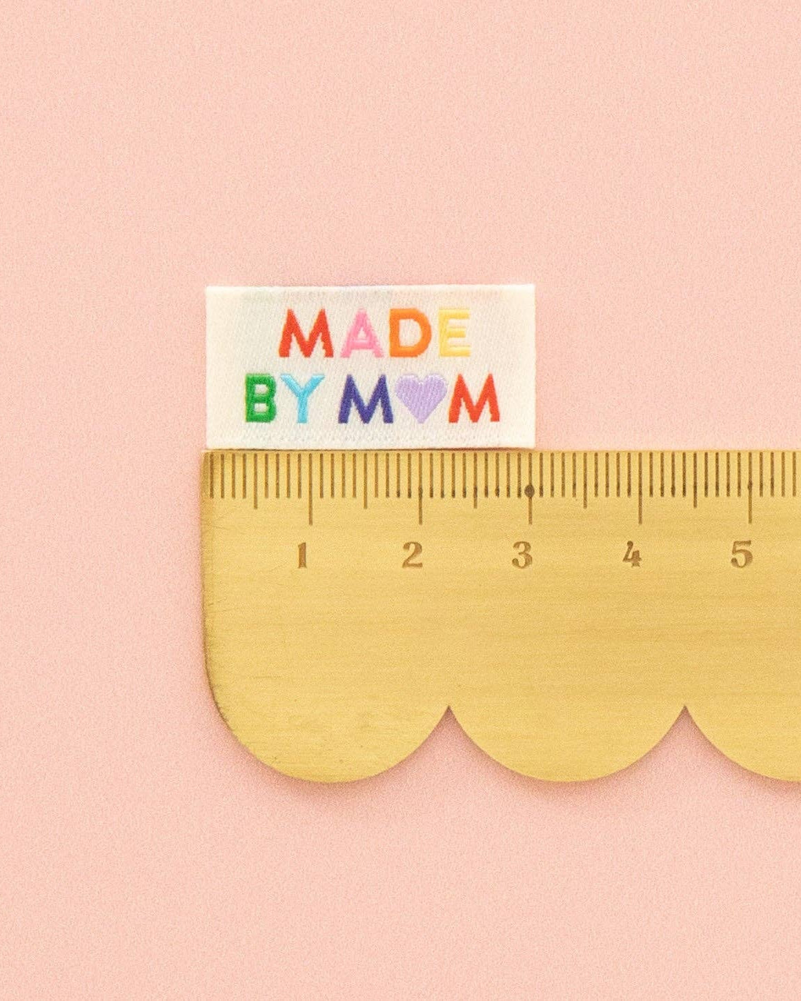 A small rectangular white sew-in label reading “MADE BY MOM” in colorful letters sits above a gold scalloped ruler on a soft pink background.