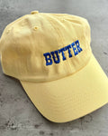 A pale yellow dad-style baseball cap with the word 'BUTTER' embroidered in blue block letters on the front, shown against a solid gray background.