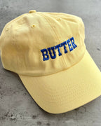 A pale yellow dad-style baseball cap with the word 'BUTTER' embroidered in blue block letters on the front, shown against a solid gray background.