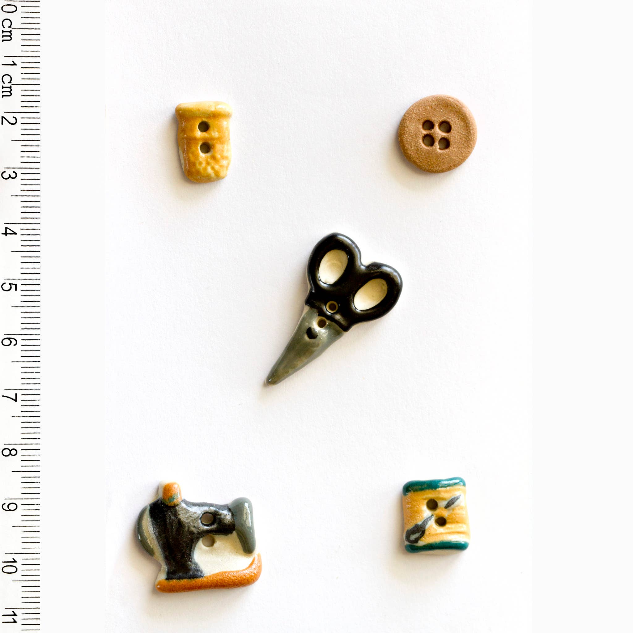 Set of ceramic sewing-themed buttons including scissors, a sewing machine, and buttons on a white background with a ruler for scale.