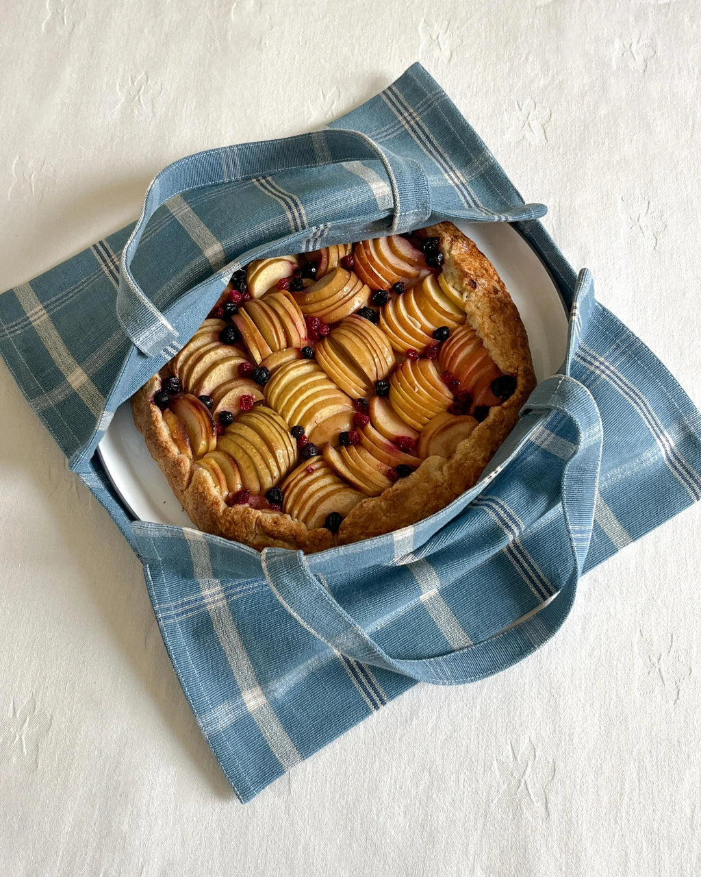 A blue and white plaid fabric pie carrier with handles, shown from above on a white textured surface, holding a round fruit tart on a white plate.