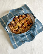 A blue and white plaid fabric pie carrier with handles, shown from above on a white textured surface, holding a round fruit tart on a white plate.