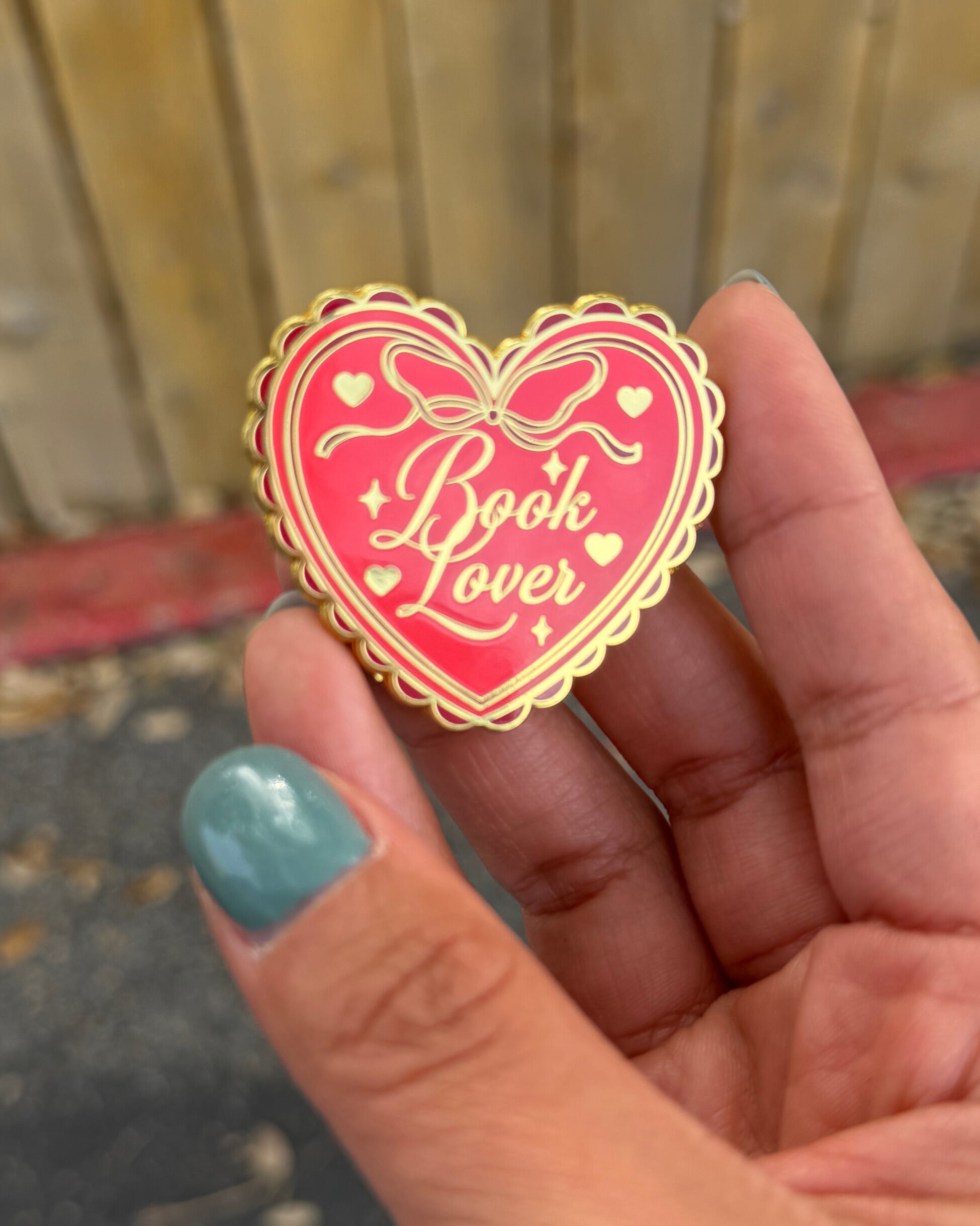 Heart-shaped 'Book Lover' pin held in a hand with teal nail polish.