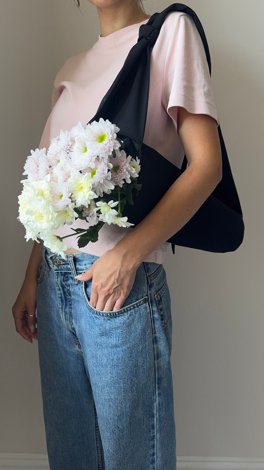 A person wearing loose-fit blue jeans and a pale pink top stands indoors against a white wall, holding a black flower tote with a bouquet of light pink and white flowers in it.