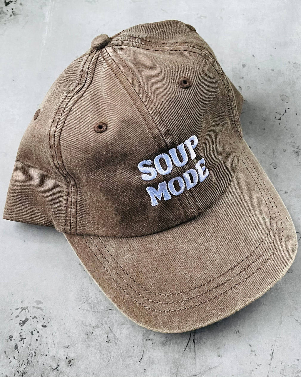 A washed brown baseball cap with 'SOUP MODE' embroidered in white and light blue on the front, resting on a textured gray concrete surface.