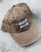 A washed brown baseball cap with 'SOUP MODE' embroidered in white and light blue on the front, resting on a textured gray concrete surface.