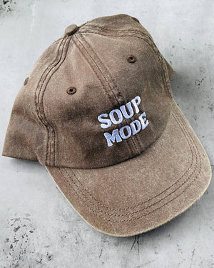 A washed brown baseball cap with 'SOUP MODE' embroidered in white and light blue on the front, resting on a textured gray concrete surface.