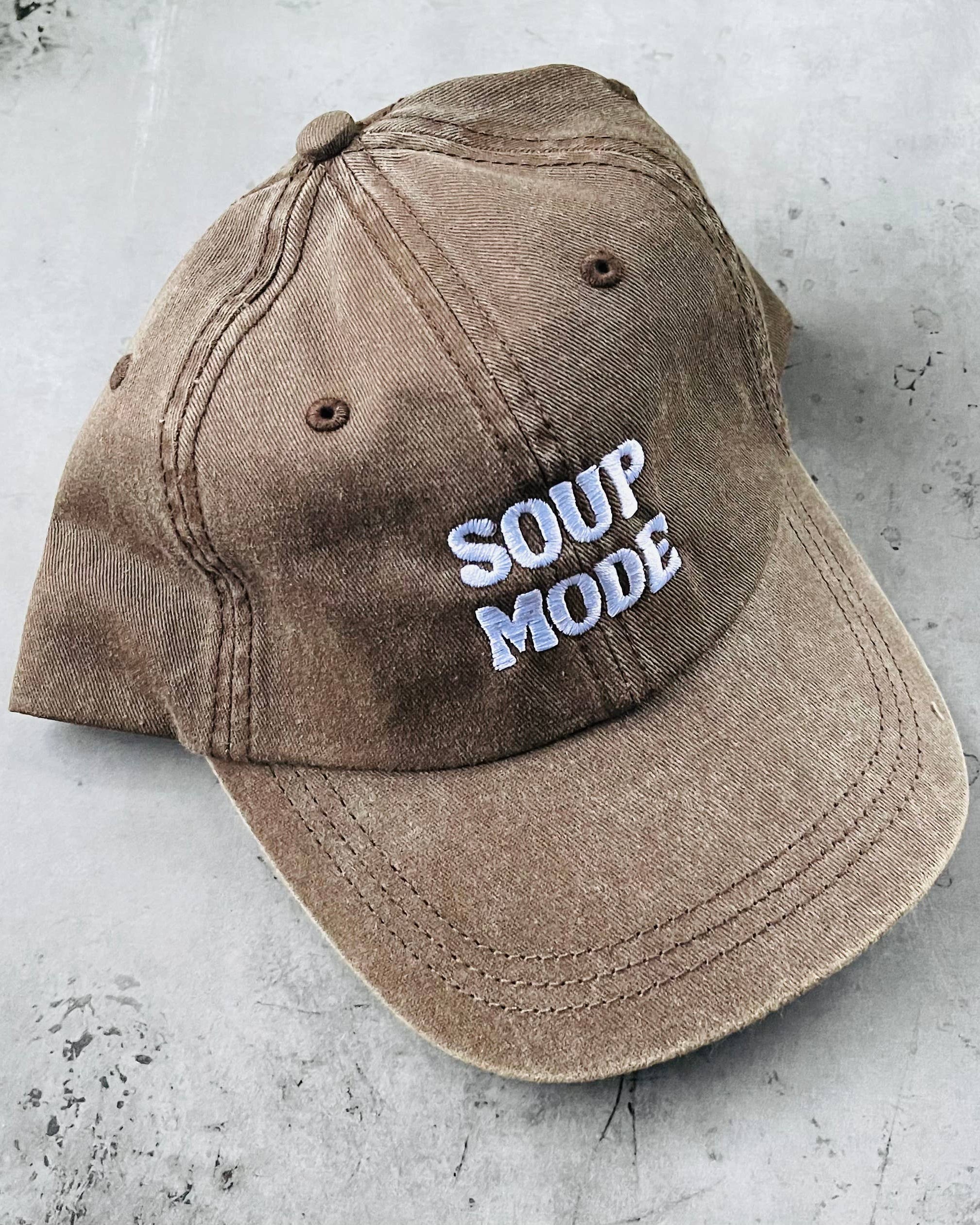 A washed brown baseball cap with 'SOUP MODE' embroidered in white and light blue on the front, resting on a textured gray concrete surface.