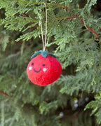A red felt tomato Christmas ornament with a smiling face hangs from a string among green evergreen branches.