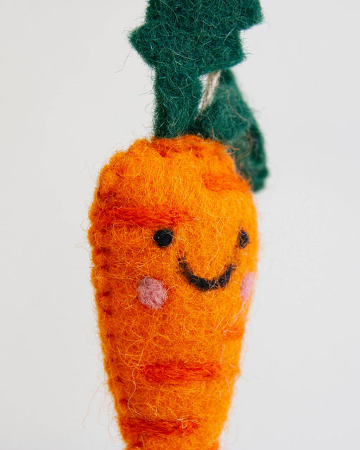 A close-up of a handmade orange felt carrot ornament with a green felt leaf top and a smiling embroidered face, set against a plain white background.