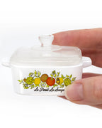 A hand is holding a white ceramic Dutch oven inspired trinket box with a colorful fruit illustration and the brand name 'Le Petit La Bouche' printed on the front, set against a plain white background.