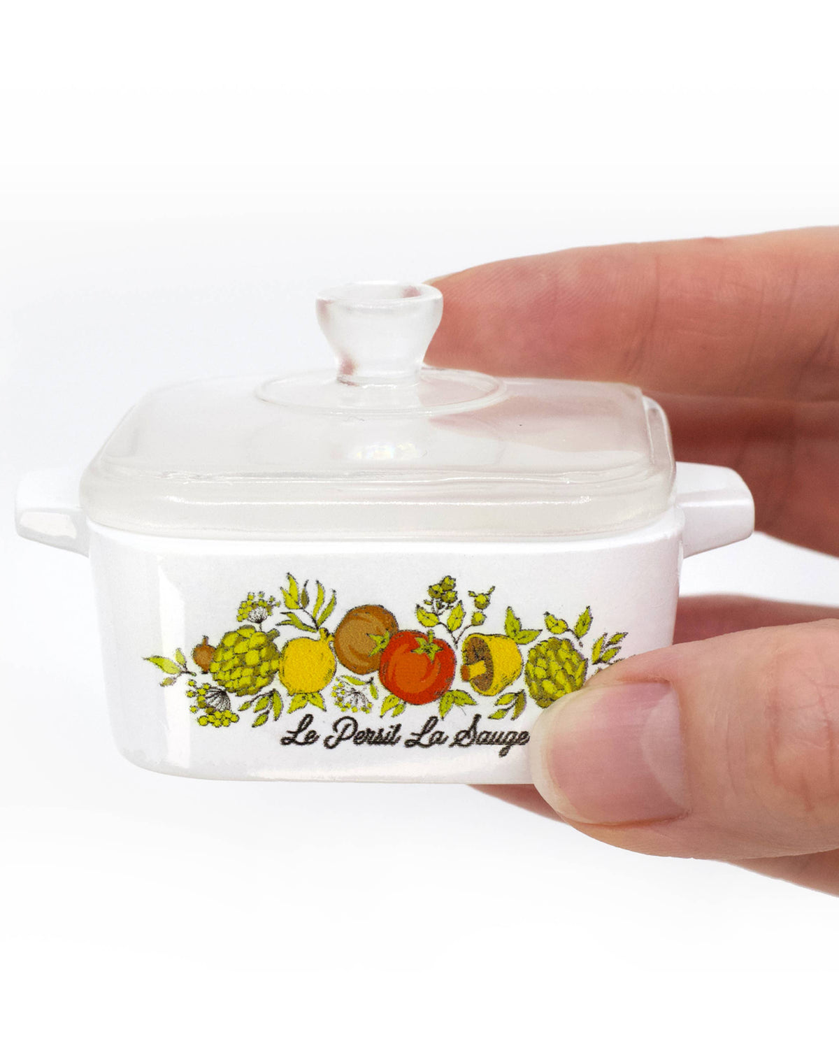 A hand is holding a white ceramic Dutch oven inspired trinket box with a colorful fruit illustration and the brand name 'Le Petit La Bouche' printed on the front, set against a plain white background.