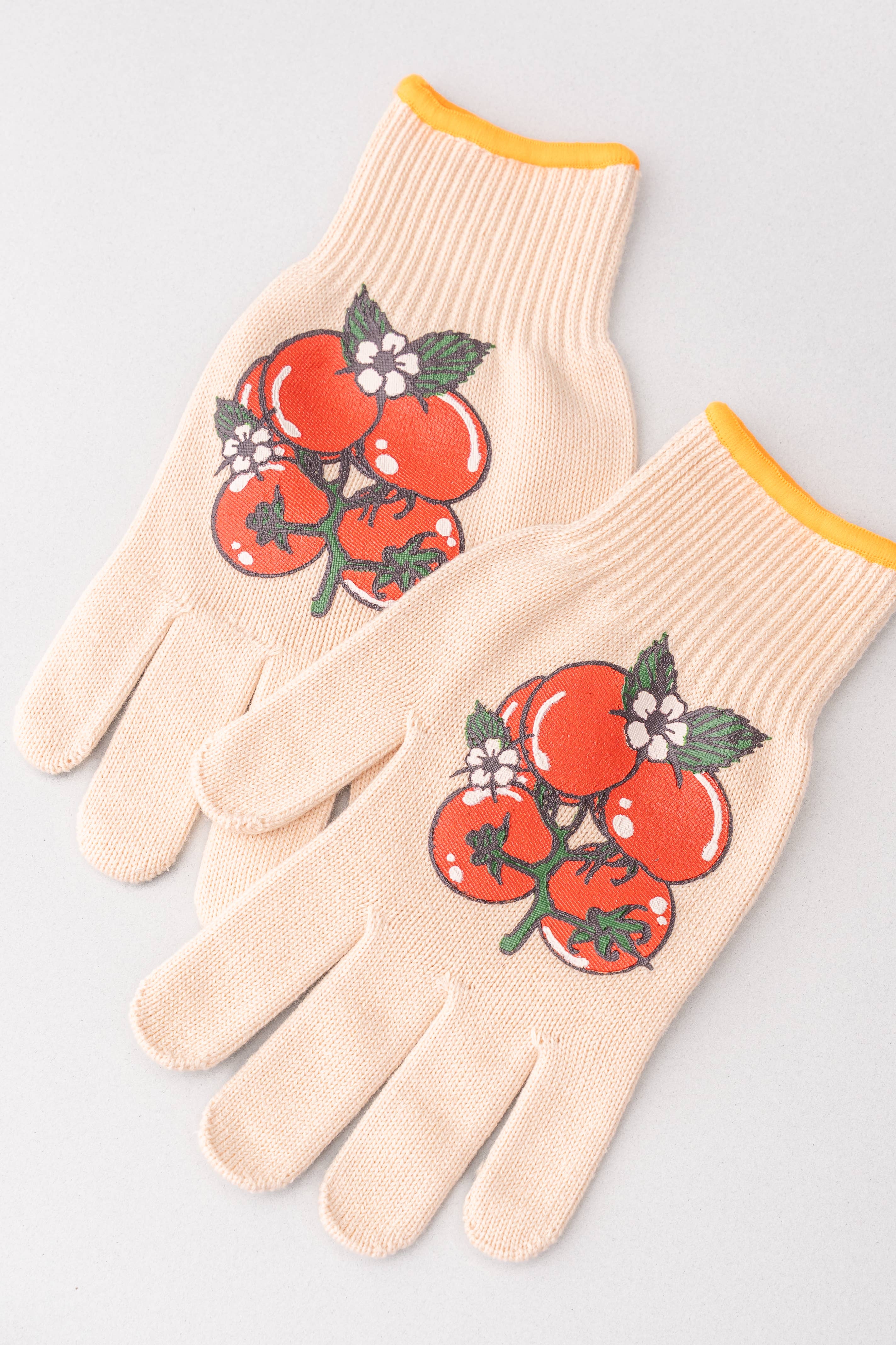 A pair of cream-colored gardening gloves featuring a graphic print of red tomatoes with green leaves and small white flowers on the back of the hands, finished with a yellow ribbed cuff.
