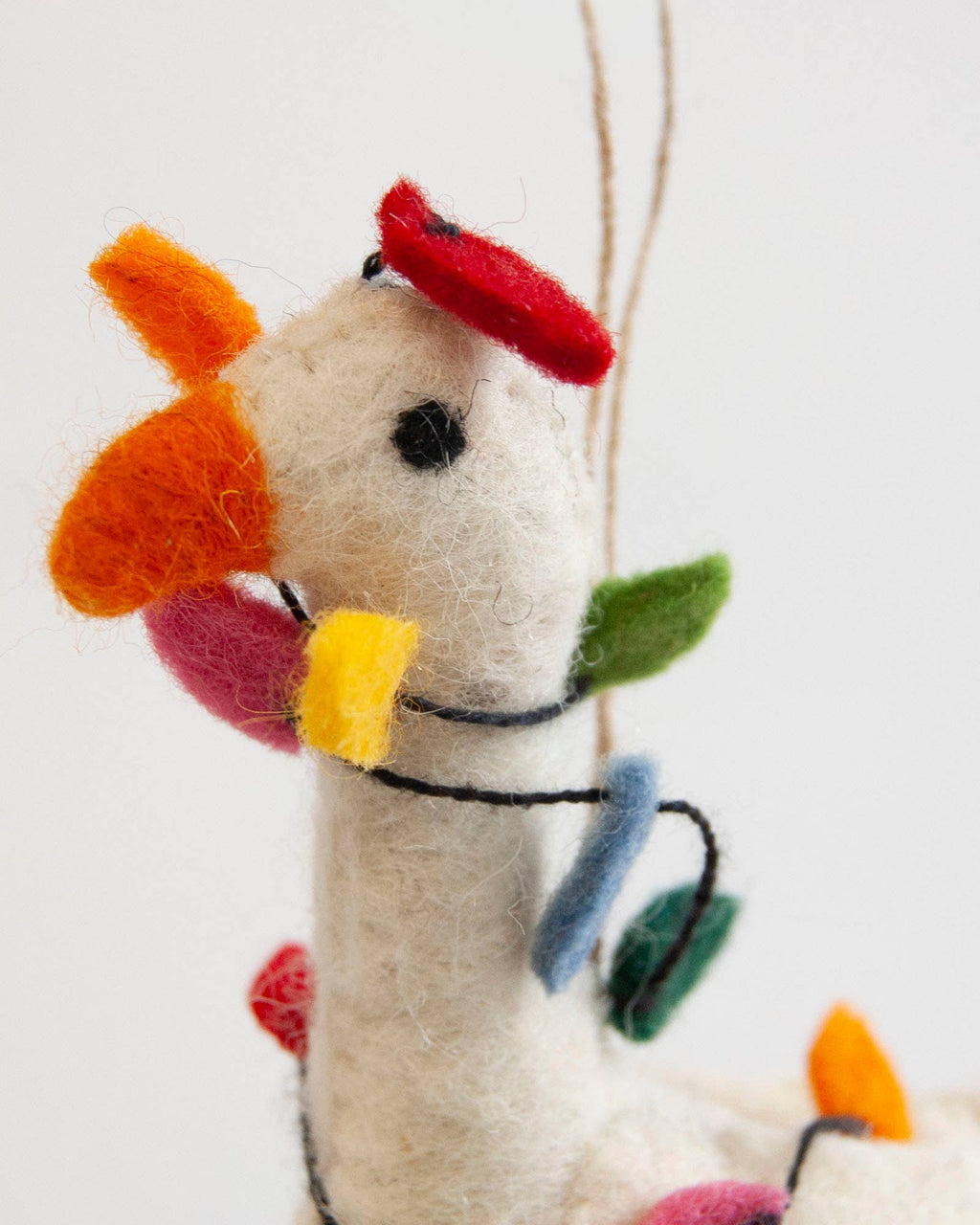 A close-up of a handcrafted felt Christmas ornament shaped like a llama or alpaca, featuring a cream-colored body, colorful felt accents in red, orange, yellow, green, pink, and blue, and a thin jute hanging string against a plain white background.