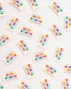 Multiple small rectangular sew-in labels scattered on a white background, each printed with the phrase “MADE BY MOM” in rainbow-colored letters.