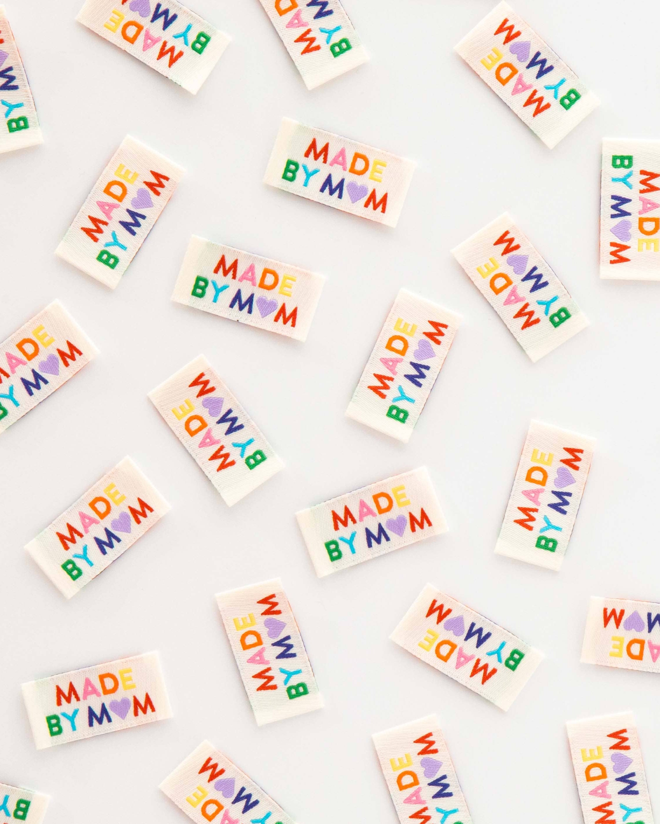 Multiple small rectangular sew-in labels scattered on a white background, each printed with the phrase “MADE BY MOM” in rainbow-colored letters.