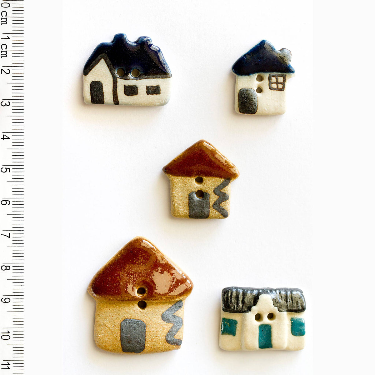 Five ceramic house-shaped buttons with different colors and designs on a white background, with a ruler for scale.