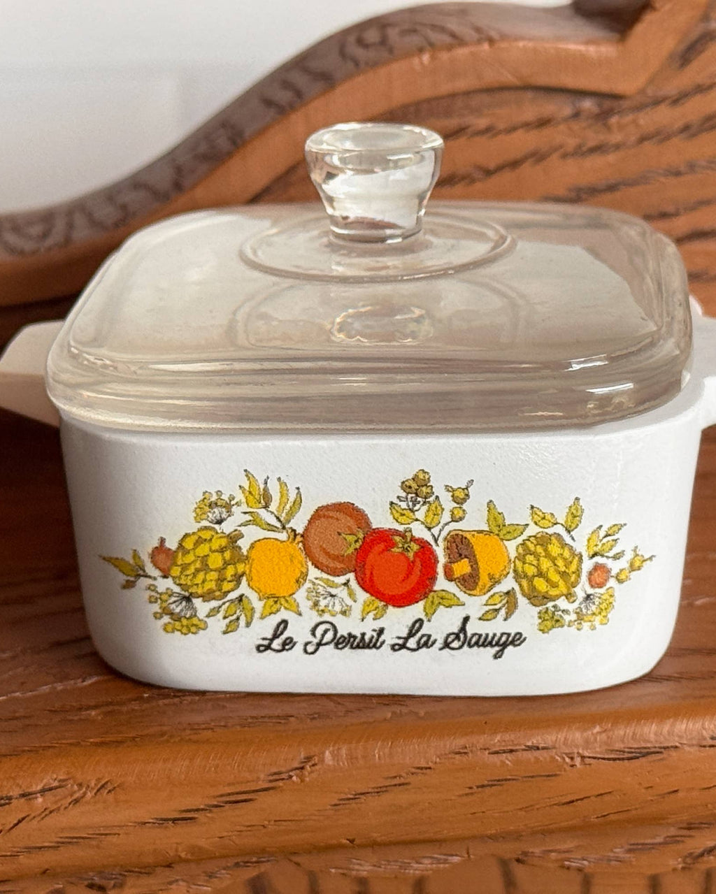 A white ceramic casserole dish inspired trinket box with a clear glass lid, featuring a colorful fruit illustration and the French text 'Le Persil Le Sauge' on the front, set on a wooden surface.